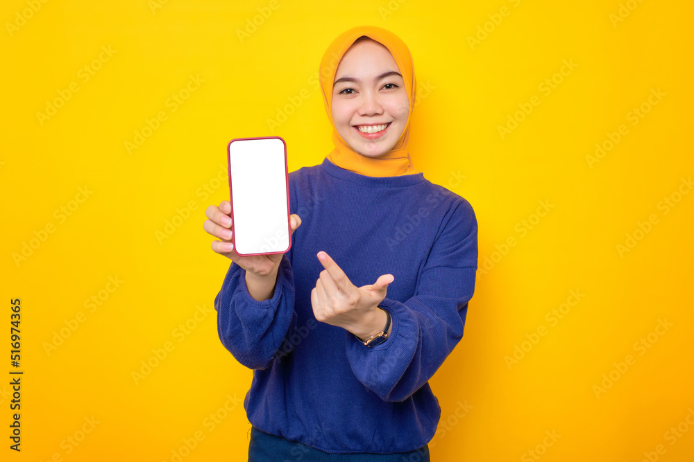 Excited young Asian Muslim woman dressed in casual sweater showing blank screen mobile phone, recommending mobile app isolated over yellow background Excited young Asian Muslim woman dressed in casual sweater showing blank screen mobile phone, recommending mobile app isolated over yellow background