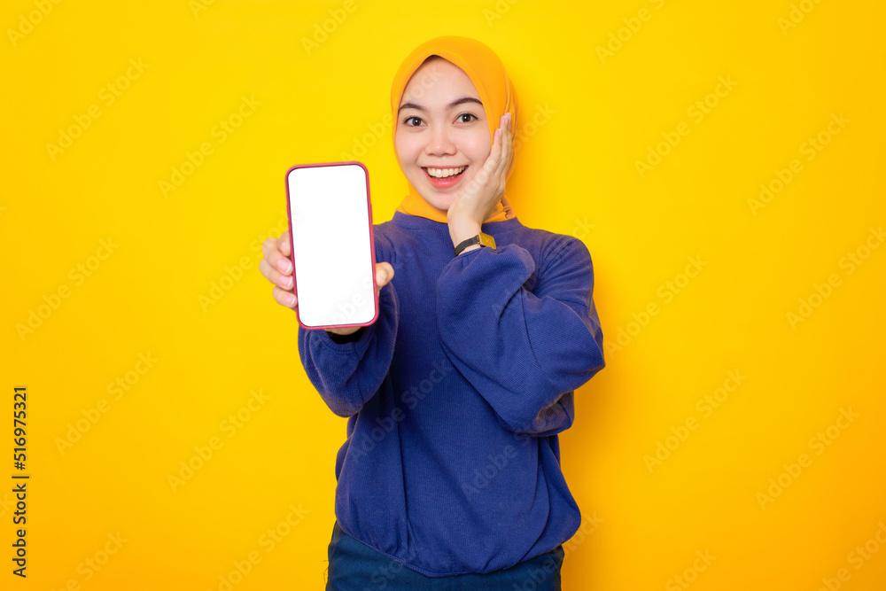 Smiling young Asian Muslim woman dressed in casual sweater showing blank screen mobile phone, recommending mobile app isolated over yellow background Smiling young Asian Muslim woman dressed in casual sweater showing blank screen mobile phone, recommending mobile app isolated over yellow background