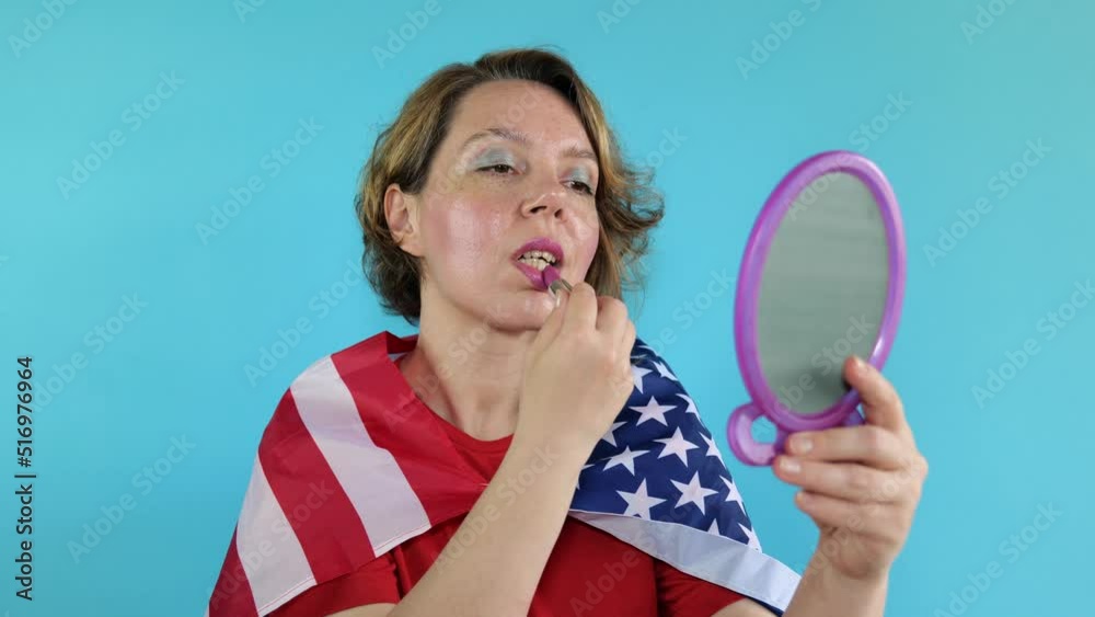 A woman in a red t-shirt with the US flag on a blue background. An American woman paints her lips looking in the mirror. Portrait of an adult cute woman A woman in a red t-shirt with the US flag on a blue background. An American woman paints her lips looking in the mirror. Portrait of an adult cute woman