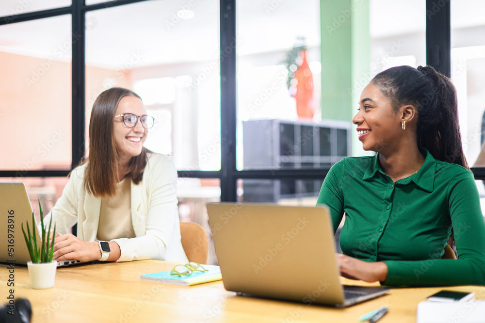 Two female colleague has pleasant conversation sitting side by side in modern open space office, smiling and enjoying friendly working atmosphere Two female colleague has pleasant conversation sitting side by side in modern open space office, smiling and enjoying friendly working atmosphere