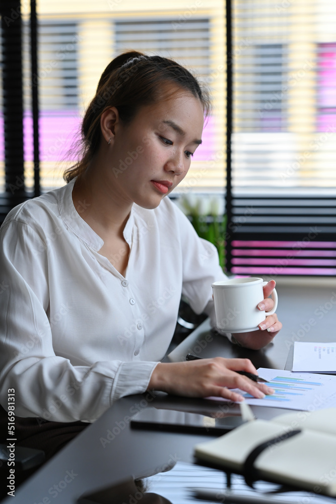 Portrait of Asian Businesswoman sitting in the office and holding a cup of coffee while checking financial report on paper document. Portrait of Asian Businesswoman sitting in the office and holding a cup of coffee while checking financial report on paper document.