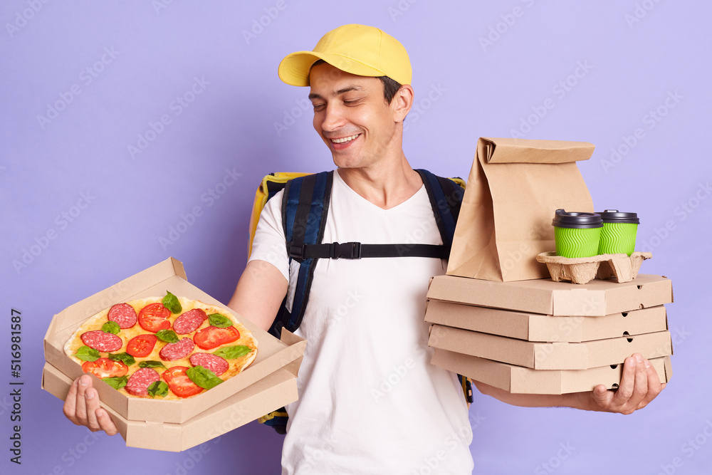 Indoor shot of smiling delivery man wearing white T-shirt with thermo backpack holding pizza boxes and coffee in disposable cups isolated over purple background, showing order to client. Indoor shot of smiling delivery man wearing white T-shirt with thermo backpack holding pizza boxes and coffee in disposable cups isolated over purple background, showing order to client.