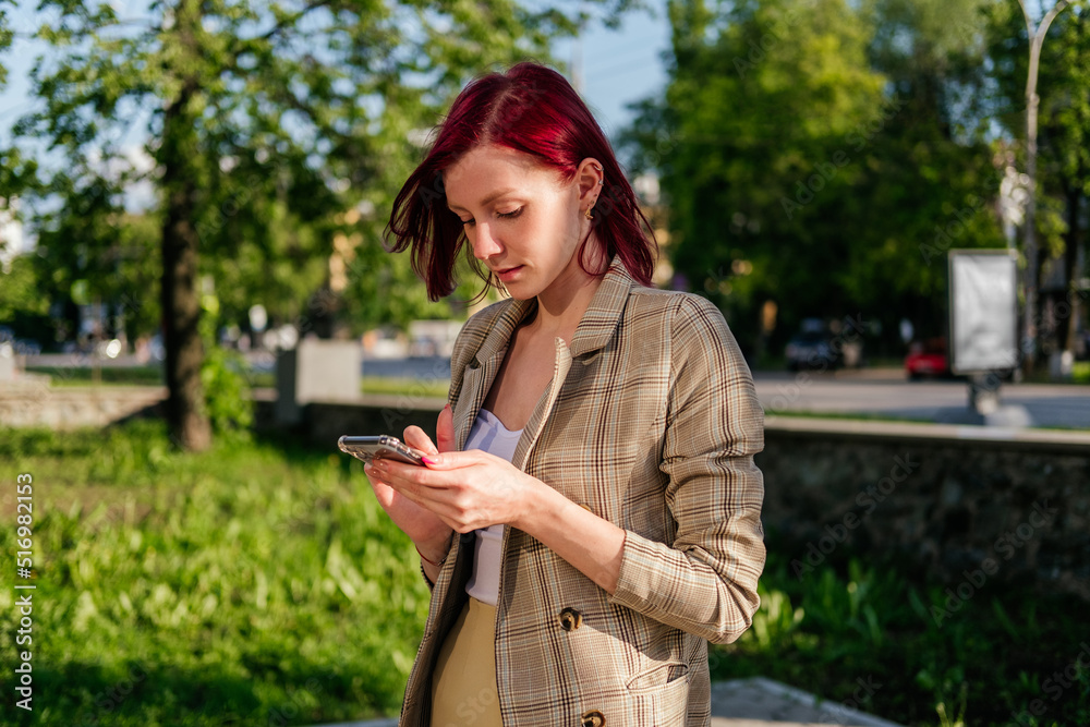 Woman with red hair standing with smartphone in hands outdoor. Girl using smartphone Woman with red hair standing with smartphone in hands outdoor. Girl using smartphone