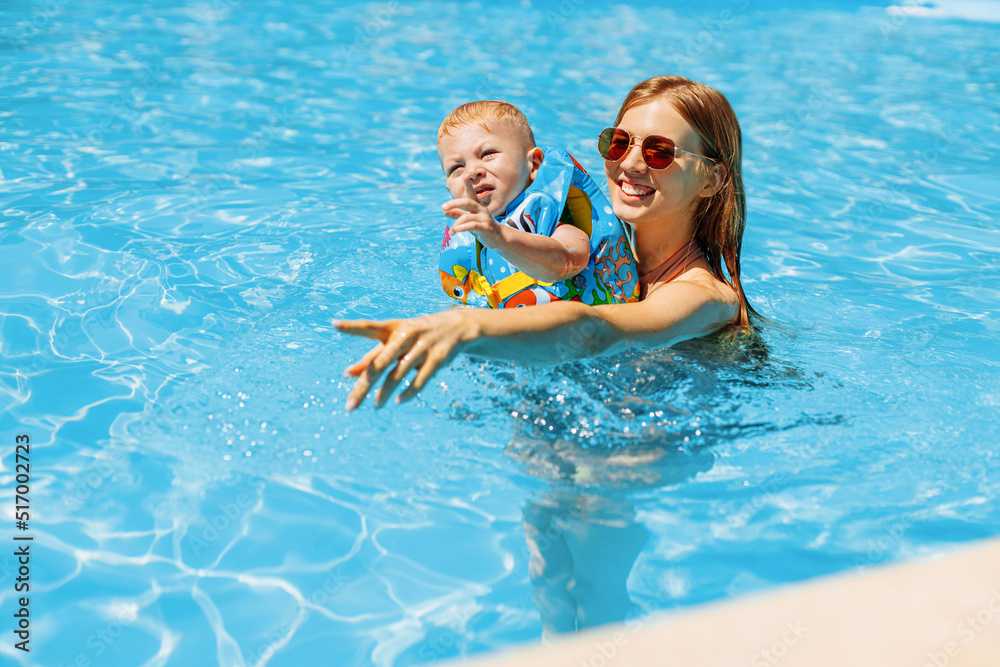 Happy young mother plays with her baby in the outdoor pool on a hot summer day. Children learn to swim on a family vacation. A young family with a child has fun in the pool on a summer day Happy young mother plays with her baby in the outdoor pool on a hot summer day. Children learn to swim on a family vacation. A young family with a child has fun in the pool on a summer day