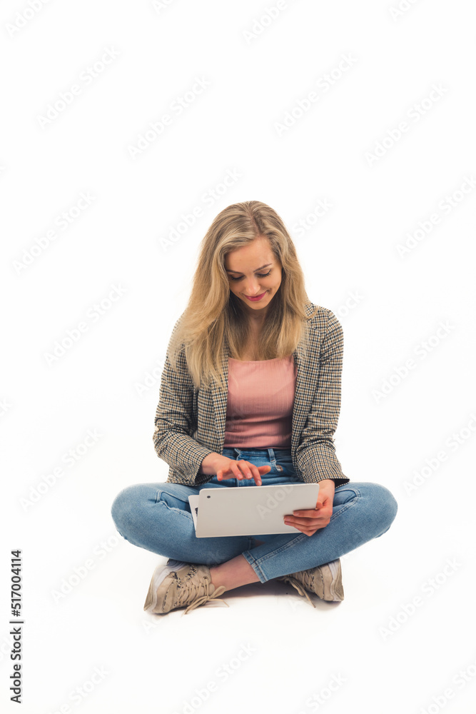 Vertical studio full body shot with white background of a cute blond caucasian woman in jeans and checked blazer sitting cross-legged and typing on laptop. . High quality photo Vertical studio full body shot with white background of a cute blond caucasian woman in jeans and checked blazer sitting cross-legged and typing on laptop. . High quality photo