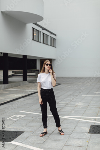 Young woman in white t-shirt standing in front of grey building and using smartphone. Urban style and street fashion. Girl in sunglasses with phone. Chatting, messaging, reading