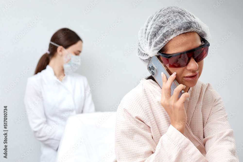 caucasian woman at laser epilation procedure, use smartphone while getting hair removal procedure in beauty salon, wearing protective eyeglasses. hairless, hair removal, epilation, depilation concept caucasian woman at laser epilation procedure, use smartphone while getting hair removal procedure in beauty salon, wearing protective eyeglasses. hairless, hair removal, epilation, depilation concept