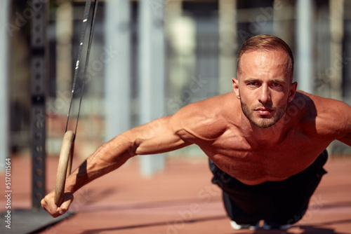 Fototapeta Naklejka Na Ścianę i Meble -  Handsome man on sport ground. The sportsman the guy, carries out difficult exercise, sports gymnastics. Muscular man doing workout with rings. The concept of a modern young man