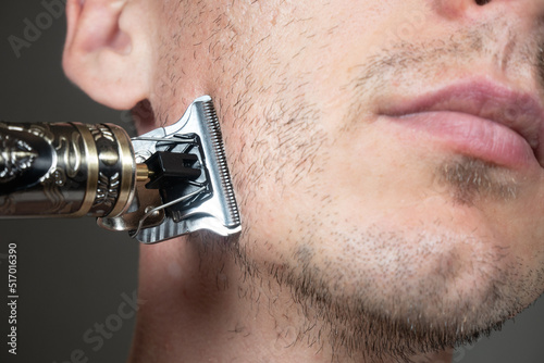 Man is cutting beard by Electric metal razor or dry shaver. Trimmer for cutting. Short, sparse beard on mans face. Hair growth problems. Unshaven bristles on the beard.