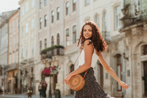 Happy smiling curly brunette woman wearing trendy summer outfit with round wicker shoulder rotan bag, white top, polka dot midi skirt, walking in street of European city. Fashion, lifestyle concept
