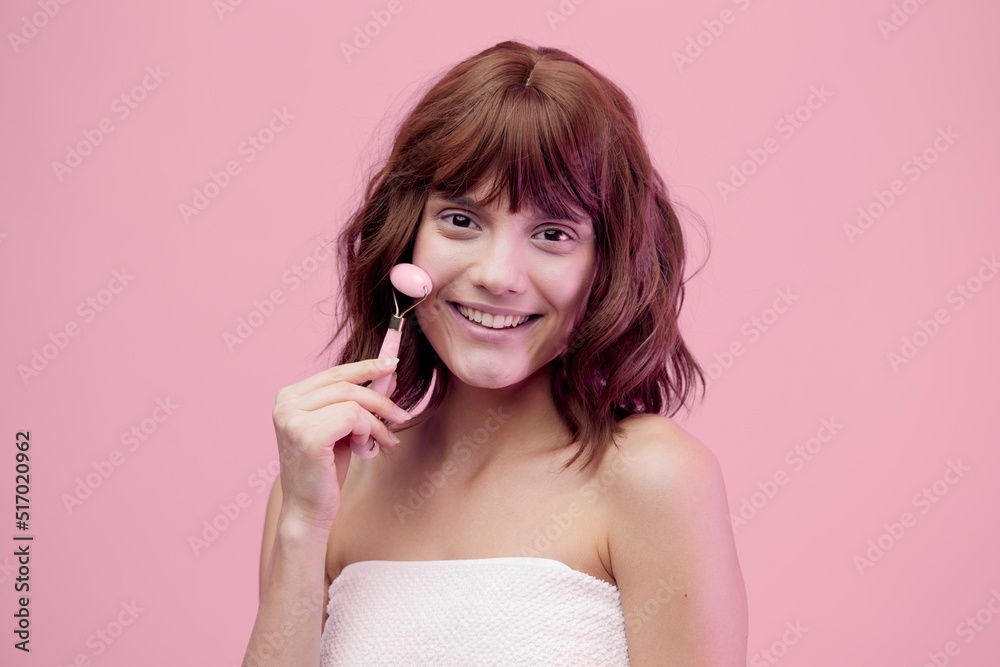 sweet, happy woman standing on a pink background with pink lighting from the side, with beautiful, styled hair, doing a facial massage with a pink roller. Horizontal photo with empty space sweet, happy woman standing on a pink background with pink lighting from the side, with beautiful, styled hair, doing a facial massage with a pink roller. Horizontal photo with empty space