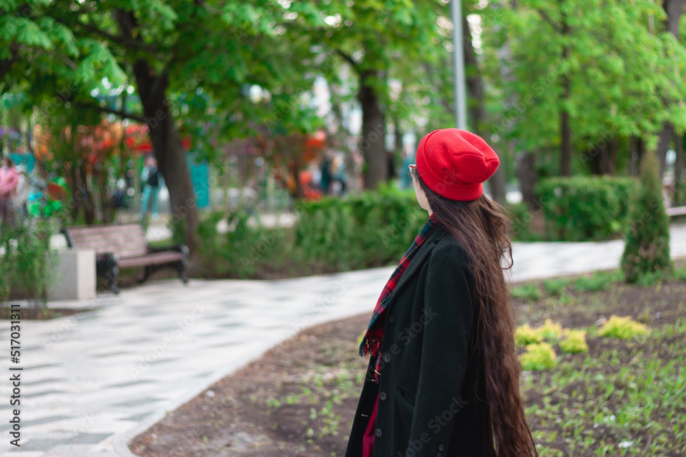 A young woman with long hair and a red hat wearing glasses and dark coat with a red scarf in the park among the trees. A pedestrian road with a bench is in the background