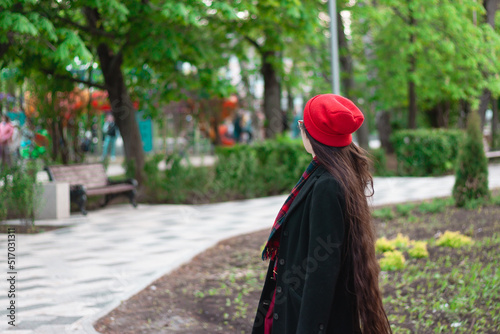 A young woman with long hair and a red hat wearing glasses and dark coat with a red scarf in the park among the trees. A pedestrian road with a bench is in the background
