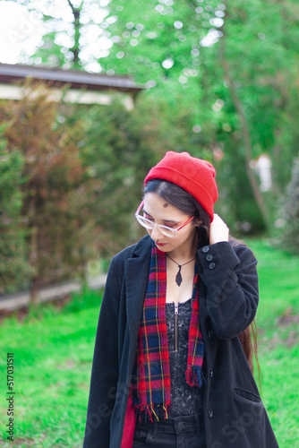 A young woman with long hair and a red hat wearing glasses and dark coat with a red scarf in the park standing against trees on a pedestrian road.