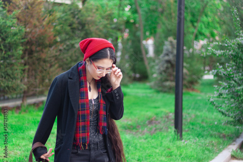 A young woman with long hair and a red hat wearing glasses and dark coat with a red scarf in the park standing against trees on a pedestrian road.