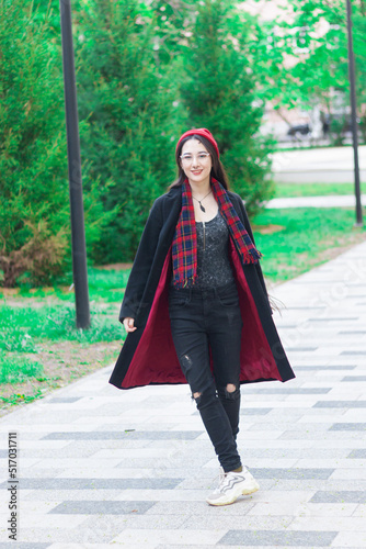 A young woman with long hair and a red hat wearing glasses and dark coat with a red scarf in the park walking against trees on a pedestrian road. © Norgle
