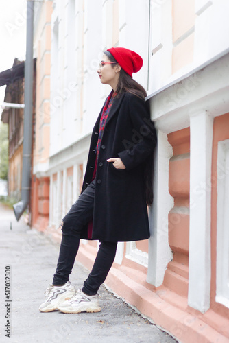A young woman wearing a red hat, red scarf and a black coat is leaning on the white and orange classic building at the street