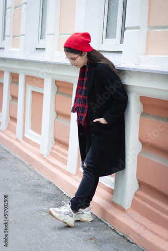 A young woman wearing a red hat, red scarf and a black coat is leaning on the white and orange classic building at the street