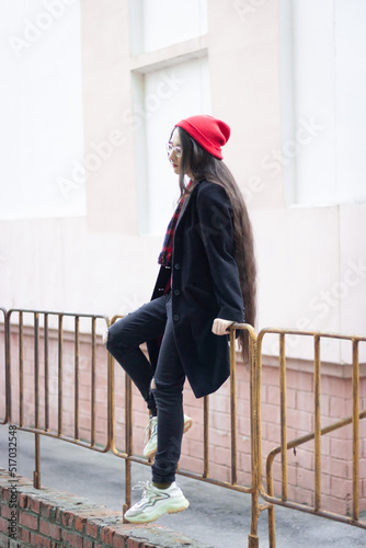A young woman wearing a red hat, red scarf and a black coat is sitting on a green fance against the white and orange classic building at the street