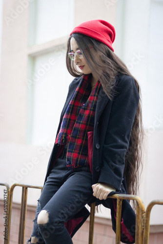 A young woman wearing a red hat, red scarf and a black coat is sitting on a green fance against the white and orange classic building at the street