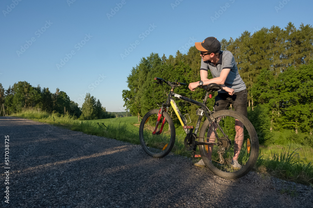 A young man with a bicycle is standing on the edge of the road. Sunny summer. Green trees. Countryside.