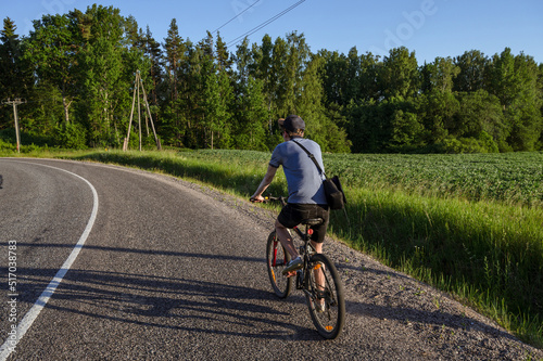A young man rides a bicycle on an asphalt road in the countryside. Green meadow and trees. Summer activity.