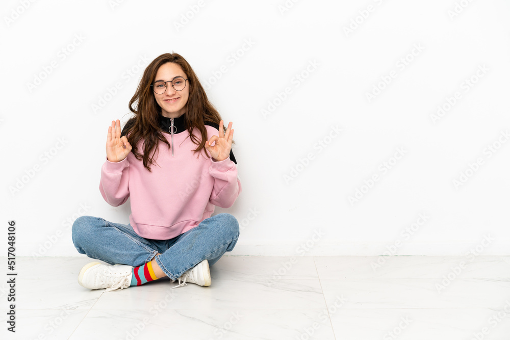 Young caucasian woman sitting on the floor isolated on white background showing an ok sign with fingers Young caucasian woman sitting on the floor isolated on white background showing an ok sign with fingers