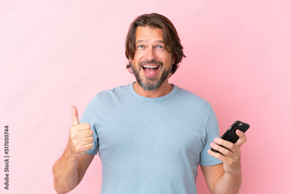 Senior dutch man isolated on pink background using mobile phone while doing thumbs up