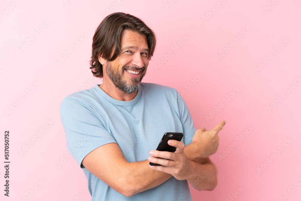 Senior dutch man isolated on pink background using mobile phone and pointing back Senior dutch man isolated on pink background using mobile phone and pointing back