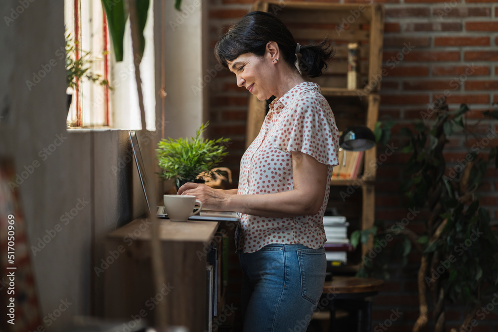 Caucasian mature woman smiling while using computer at home, having cup of coffee, having breakfast in apartment in the morning Caucasian mature woman smiling while using computer at home, having cup of coffee, having breakfast in apartment in the morning