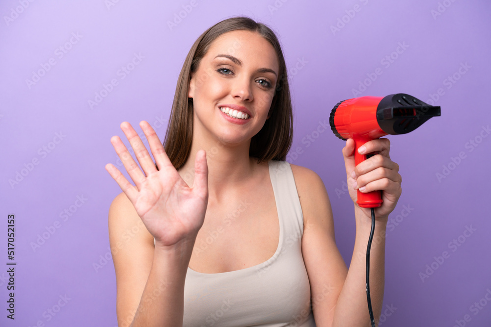 Young caucasian woman holding a hairdryer isolated on purple background saluting with hand with happy expression Young caucasian woman holding a hairdryer isolated on purple background saluting with hand with happy expression