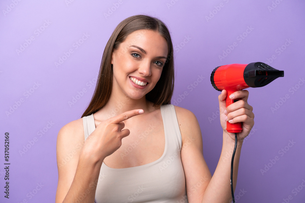 Young caucasian woman holding a hairdryer isolated on purple background and pointing it Young caucasian woman holding a hairdryer isolated on purple background and pointing it