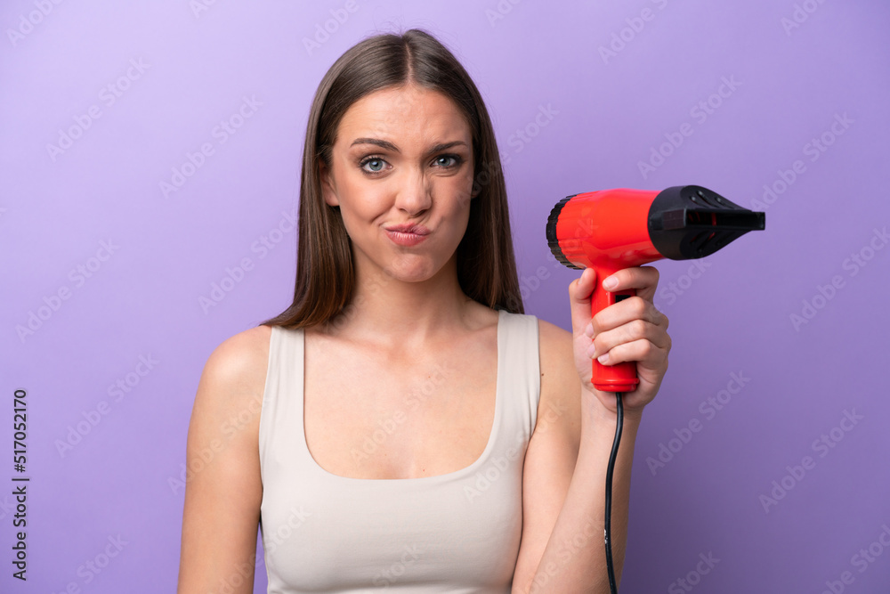 Young caucasian woman holding a hairdryer isolated on purple background with sad expression Young caucasian woman holding a hairdryer isolated on purple background with sad expression