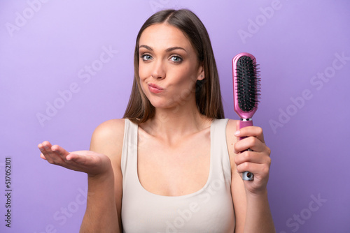 Young caucasian woman holding hairbrush isolated on purple background making doubts gesture while lifting the shoulders