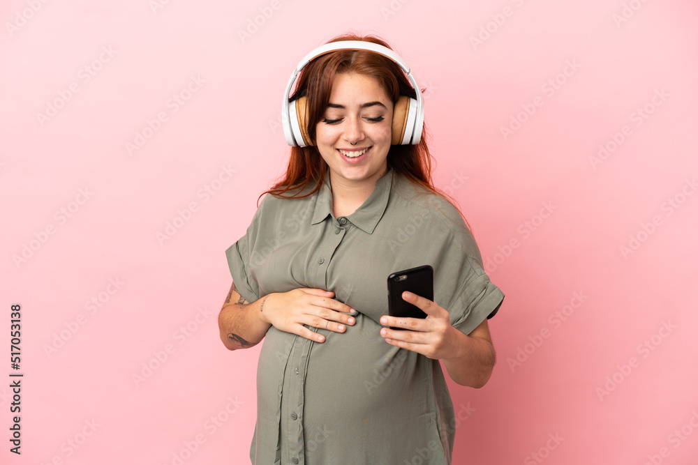 Young redhead caucasian woman isolated on pink background pregnant and listening music with a mobile Young redhead caucasian woman isolated on pink background pregnant and listening music with a mobile
