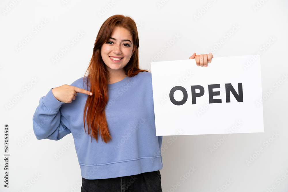 Young redhead woman isolated on white background holding a placard with text OPEN and pointing it Young redhead woman isolated on white background holding a placard with text OPEN and pointing it