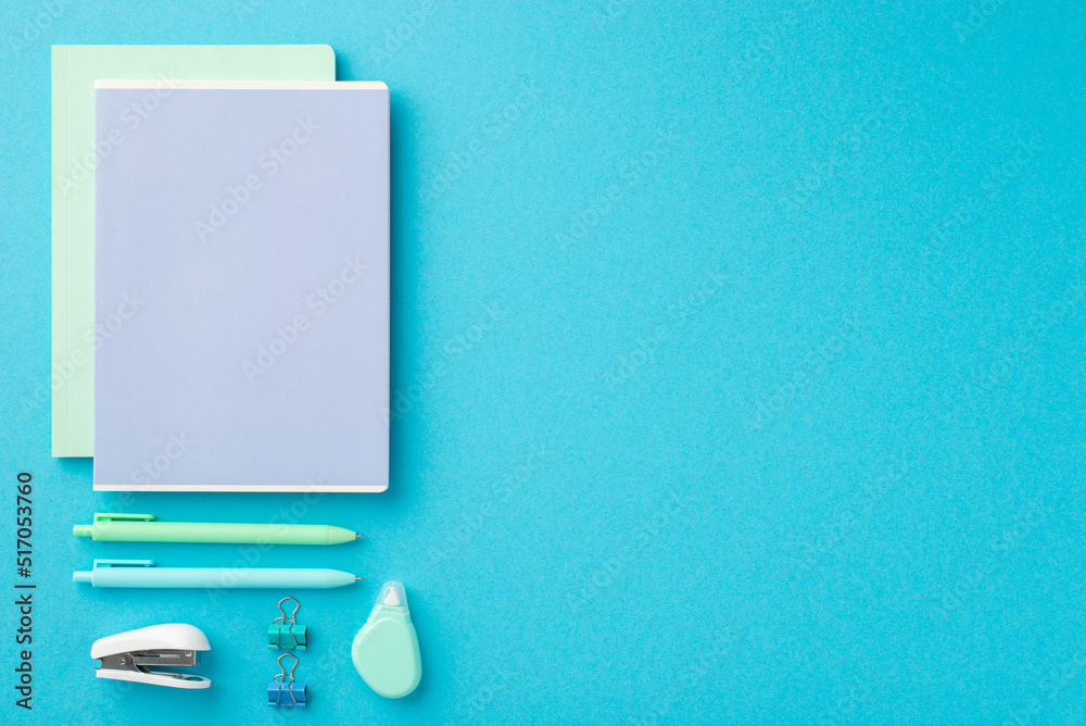 Back to school concept. Top view photo of ordered stationery composition two note pads binder clips pens stapler and round correction tape on isolated blue background with empty space Back to school concept. Top view photo of ordered stationery composition two note pads binder clips pens stapler and round correction tape on isolated blue background with empty space