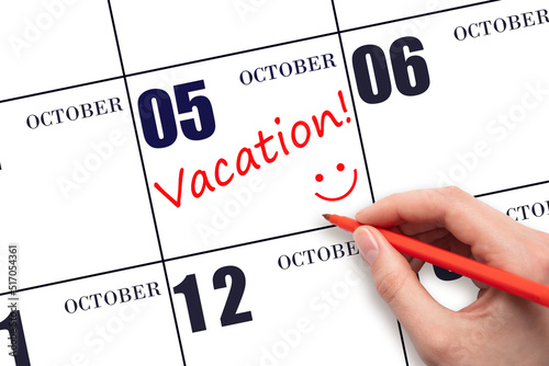 A hand writing a VACATION text and drawing a smiling face on a calendar date 5 October. Vacation planning concept.