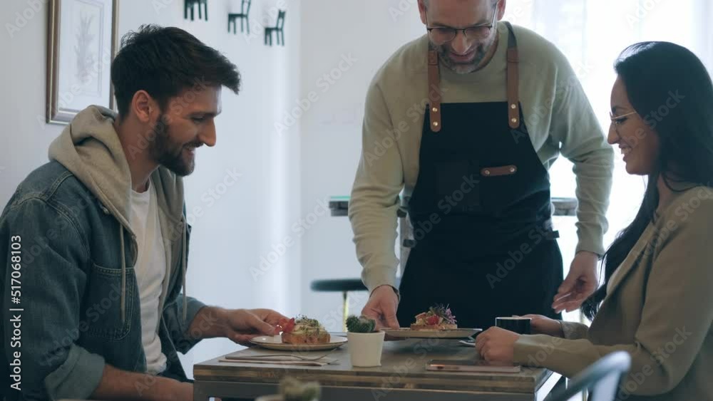 Video of handsome waitress man serving healthy and delicious toasts for smiling couple in the table in a pastry shop. Video of handsome waitress man serving healthy and delicious toasts for smiling couple in the table in a pastry shop.