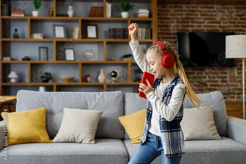 Print op canvas Happy little girl wearing red headphones dancing and singing using smartphone like microphone