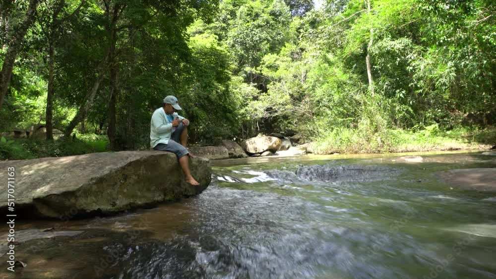 Asian Traveler sit on rock Swinging legs feet and use smartphone at Waterfall river landscape with tree forest so beautiful scenery for peaceful in spring, moisture tranquility refreshing of Thailand Asian Traveler sit on rock Swinging legs feet and use smartphone at Waterfall river landscape with tree forest so beautiful scenery for peaceful in spring, moisture tranquility refreshing of Thailand