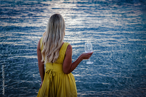 A blonde girl in a yellow dress is standing on the ocean. Glass of wine in hand. Soft focus