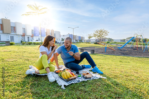 Beautiful Latin couple having a romantic breakfast with lots of tasty food and wine, sitting together on the picnic blanket in the park on a sunny morning.