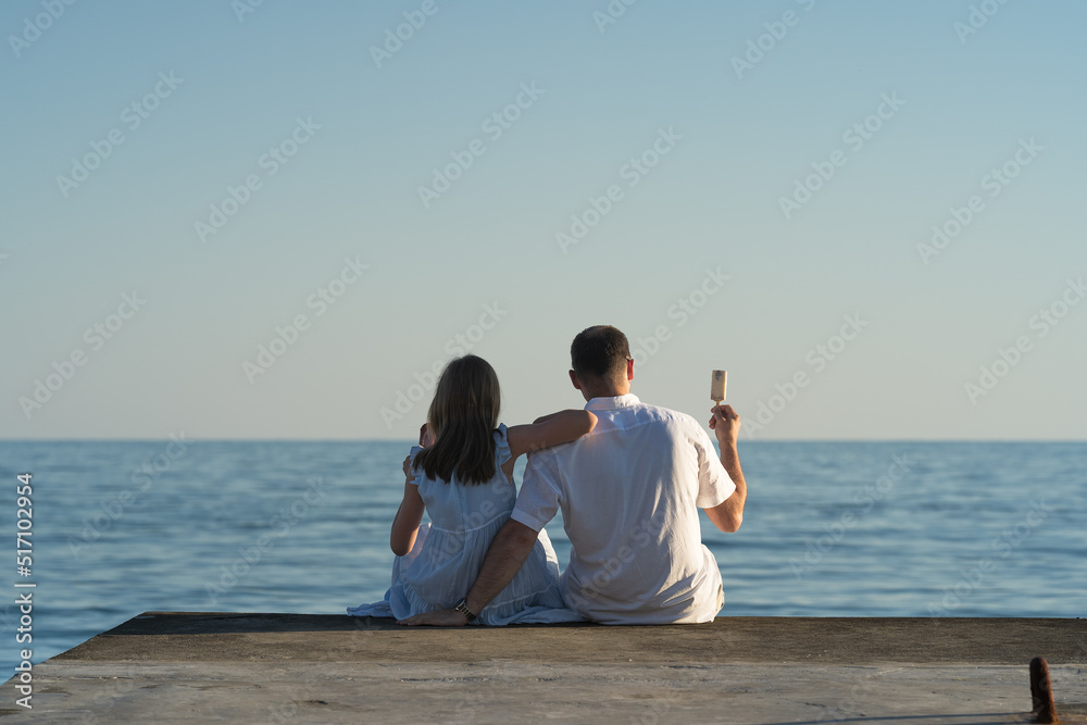 a girl and her dad are sitting on the pier and eating ice cream on a summer evening