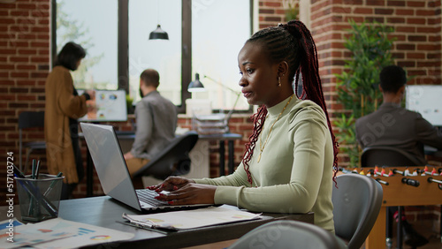 African american woman working on laptop in business office, analyzing charts on presentation to do research and paperwork. Company employee sending email to management department.