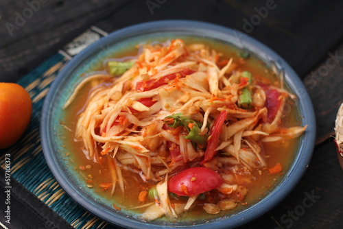 Spicy papaya salad served in a blue dish with sticky rice in a bucket and long beans, dried chilli and vegetables.