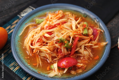 Spicy papaya salad served in a blue dish with sticky rice in a bucket and long beans, dried chilli and vegetables.