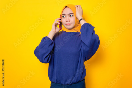 Confused young Asian Muslim woman dressed in casual sweater talking on mobile phone and touching head, having problem isolated over yellow background © Sewupari Studio