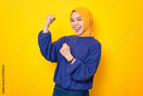 Excited young Asian Muslim woman dressed in casual sweater celebrating success with raised fist isolated over yellow background