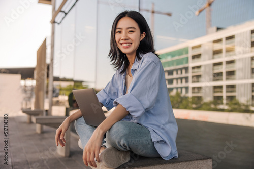 Side view of asian cheerful woman look with smile at camera in the city . Young student working outdoors sitting and using laptop. Technology, student lifestyle concept 